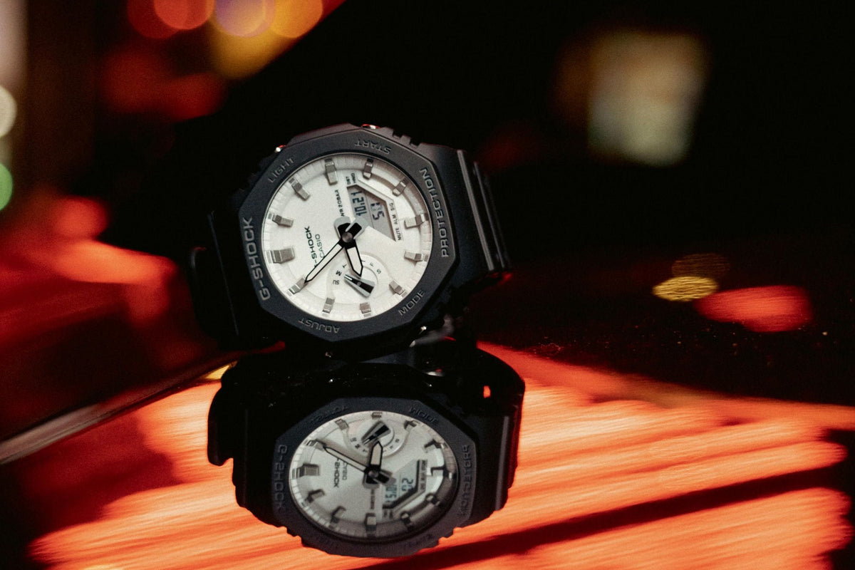 G-Shock GA-2100 de esfera blanca reflejado sobre una superficie con luces de noche
